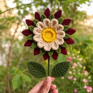 Könnte beinhalten: Eine handgefertigte Häkelblume mit einer leuchtend gelben Mitte, einem Ring aus hellbeigen Blütenblättern und mehreren Schichten spitzer, dunkelroter und waldgrüner Blütenblätter. Die Blume sitzt auf einem langen grünen Stiel mit zwei Blättern.