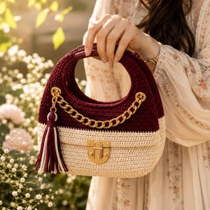 May include: A two-toned crochet handbag featuring a deep burgundy upper section and a cream-colored lower section. The bag includes a gold-tone metal chain strap, a matching gold-tone push-lock closure, and a decorative tassel in burgundy and cream.