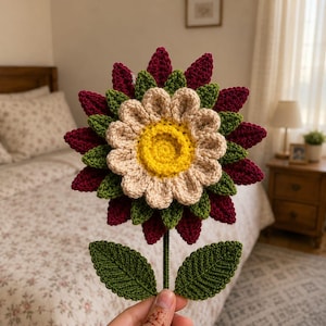 May include: A handcrafted crochet flower featuring a bright yellow centre, a layer of light beige petals, a layer of dark green leaves, and an outer layer of deep burgundy petals. The flower is attached to a green stem with two green leaves.