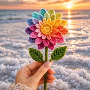 May include: A handcrafted crochet flower with vibrant rainbow petals and a yellow center. The flower is held against a backdrop of a sunset over the ocean. The stem has two green leaves.