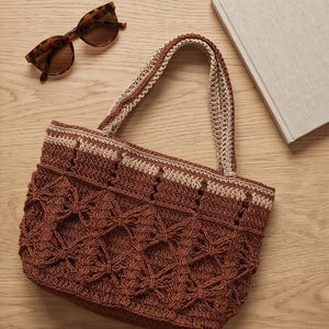 May include: A brown crochet tote bag with a textured pattern and matching handles. Tortoiseshell sunglasses and a book are next to the bag on a wooden surface. The bag is a stylish accessory.