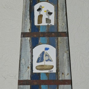 May include: A wooden lighthouse with a blue and white striped design. The lighthouse has three windows with sea-themed artwork. The top window features two seagulls, the middle window features a sailboat, and the bottom window features a wave made of sea glass. The lighthouse is on a wooden base.