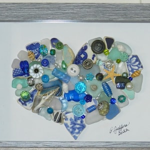 May include: A framed heart-shaped mosaic made of blue and white sea glass, shells, and buttons. The mosaic is set against a white background and is signed "S.J. Geffert 2022".