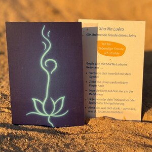 May include: A dark blue card with a glowing green lotus flower design and a second card with German text, including "Sha'Na Luera." The cards are on a sandy surface with a small, orange stone.