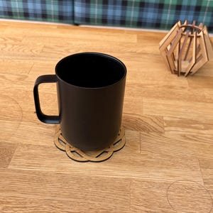 May include: A black mug with a matching black handle sits on a wooden coaster with an intricate design. A small, geometric wooden decoration is in the background. The mug and coaster are on a light wood surface.