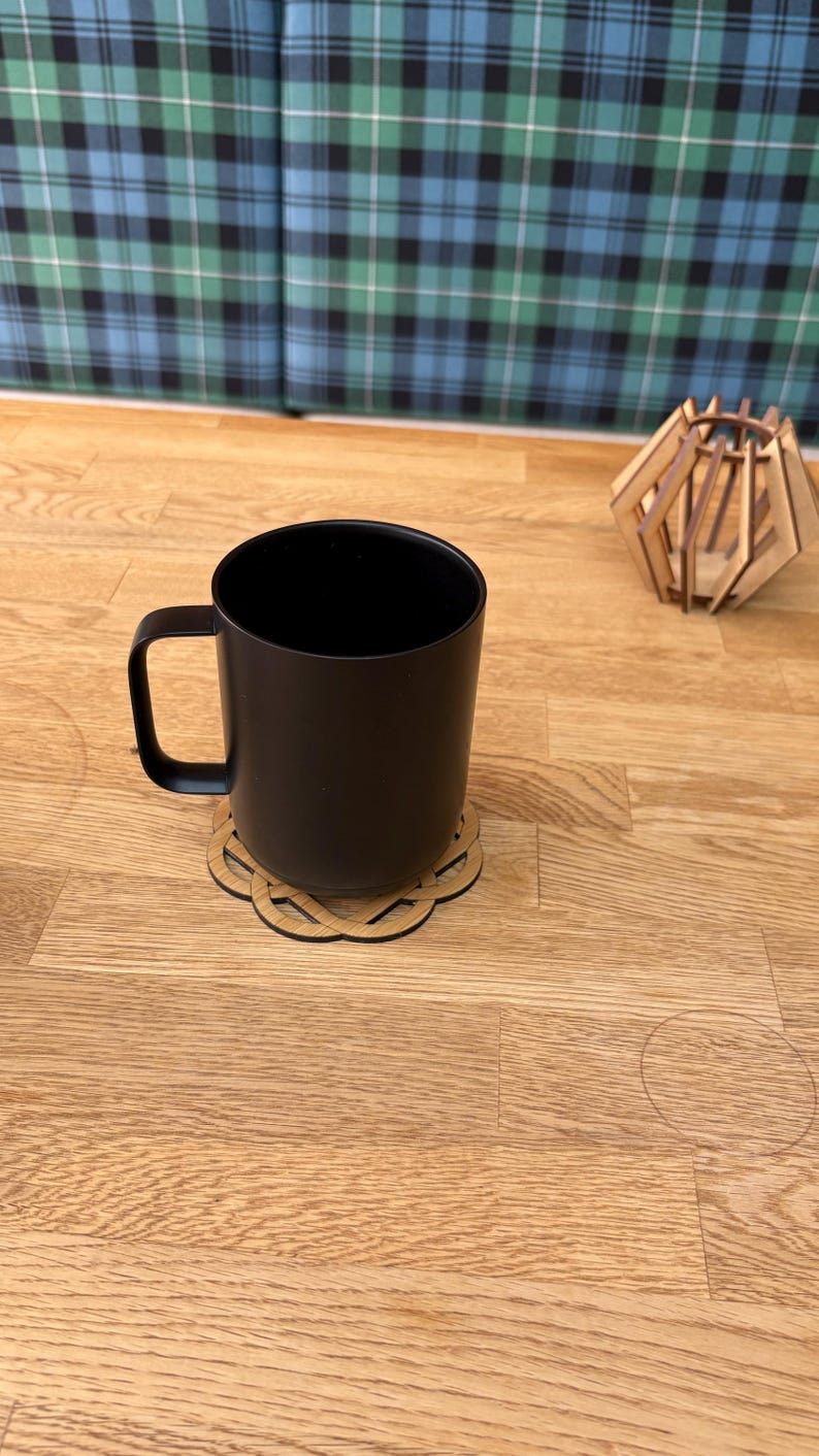 May include: A black mug with a matching black handle sits on a wooden coaster with an intricate design. A small, geometric wooden decoration is in the background. The mug and coaster are on a light wood surface.