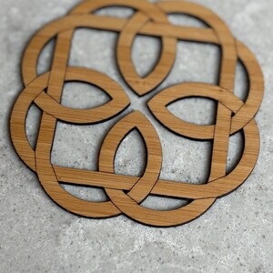 May include: A wooden coaster with a Celtic knot design. The coaster is made of light brown wood with a dark brown outline. The interlocking design is circular and symmetrical, suitable for home decor.