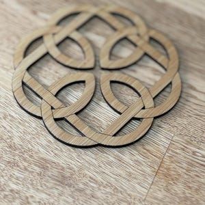 May include: A wooden coaster with a Celtic knot design. The coaster is made of light brown wood with a dark brown border. The design features an interwoven pattern of loops and curves, creating a symmetrical shape. The coaster is placed on a wooden surface.
