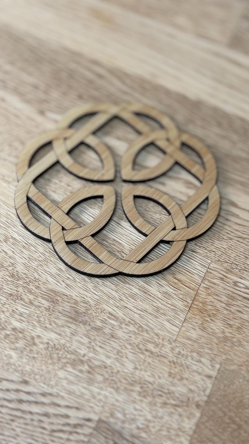 May include: A wooden coaster with a Celtic knot design. The coaster is made of light brown wood with a dark brown border. The design features an interwoven pattern of loops and curves, creating a symmetrical shape. The coaster is placed on a wooden surface.