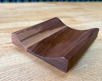 May include: A handcrafted wooden phone stand with a curved design. The stand is made of two tones of wood, with a darker brown wood on the right and a lighter brown wood on the left. The stand is on a wooden surface.