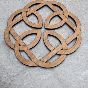 May include: A wooden coaster with a Celtic knot design. The coaster is made of light brown wood and features an intricate, interwoven pattern. The coaster is circular and sits on a gray surface. The design is a symbol of eternity and interconnectedness.