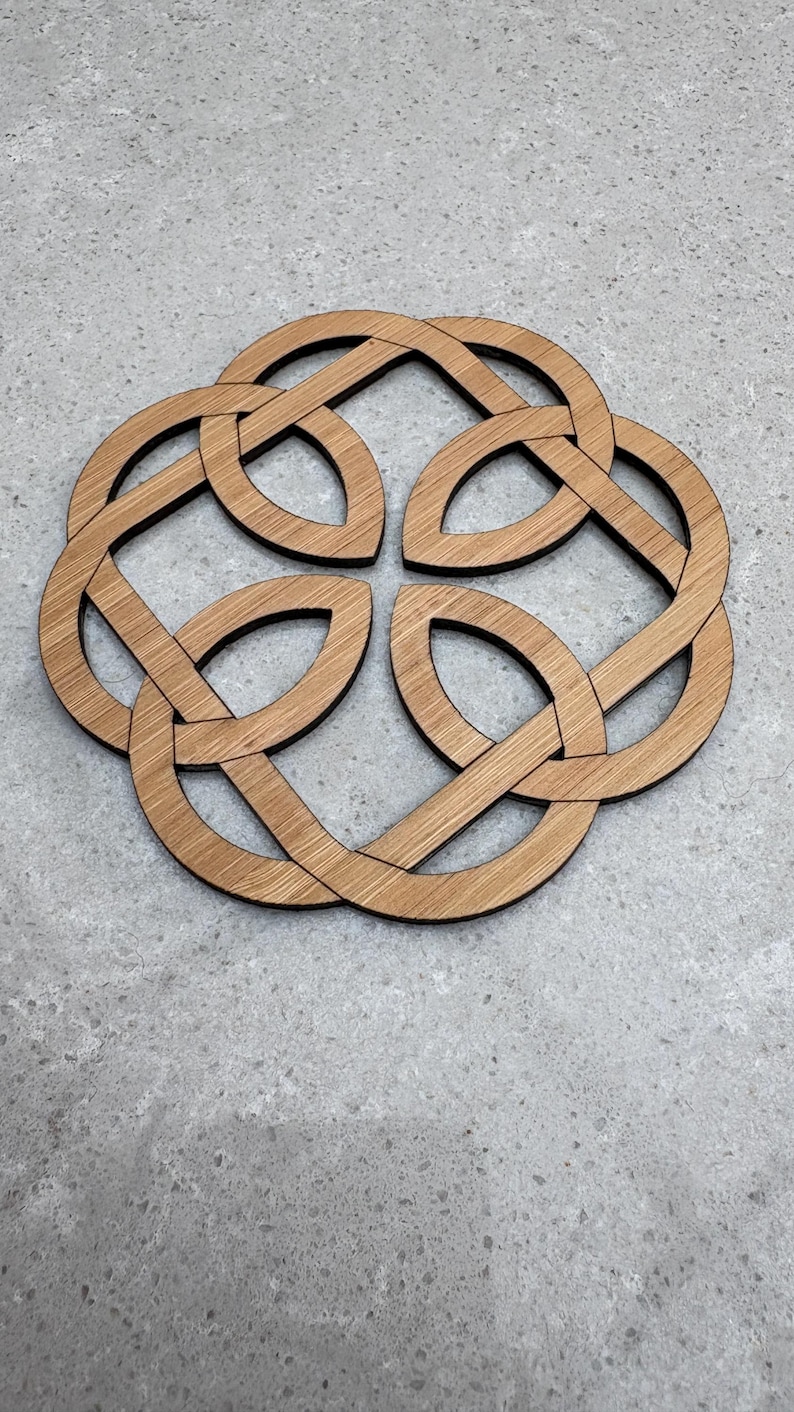 May include: A wooden coaster with a Celtic knot design. The coaster is made of light brown wood and features an intricate, interwoven pattern. The coaster is circular and sits on a gray surface. The design is a symbol of eternity and interconnectedness.