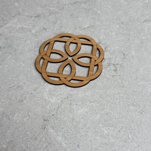 May include: A wooden coaster with a Celtic knot design. The coaster is circular with an intricate, interwoven pattern. The wood has a natural, light brown color, and the coaster sits on a light gray surface.