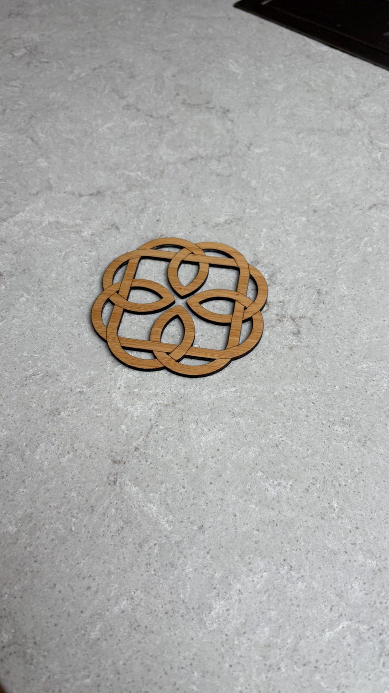May include: A wooden coaster with a Celtic knot design. The coaster is circular with an intricate, interwoven pattern. The wood has a natural, light brown color, and the coaster sits on a light gray surface.