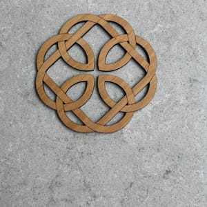 May include: A wooden coaster with a Celtic knot design. The coaster is made of light brown wood and features an intricate, interwoven pattern. The design is symmetrical and circular, with four interlocking loops. The coaster is placed on a gray surface.