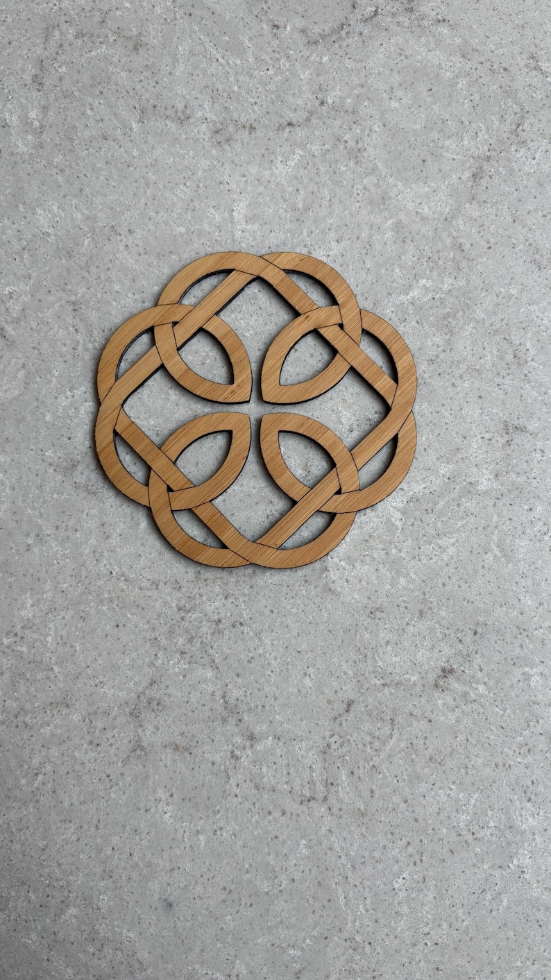 May include: A wooden coaster with a Celtic knot design. The coaster is made of light brown wood and features an intricate, interwoven pattern. The design is symmetrical and circular, with four interlocking loops. The coaster is placed on a gray surface.