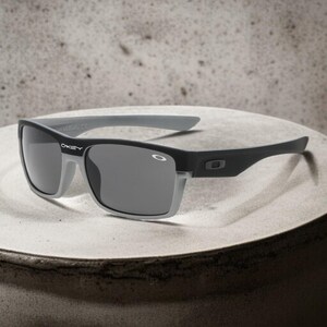 May include: Black and gray sunglasses with a rectangular frame. The sunglasses have a gray rim around the lenses and gray arms. The brand name "OAKLEY" is visible on the left arm.