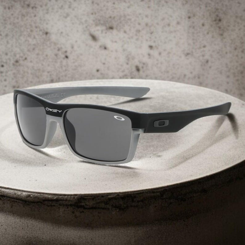 May include: Black and gray sunglasses with a rectangular frame. The sunglasses have a gray rim around the lenses and gray arms. The brand name "OAKLEY" is visible on the left arm.