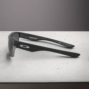 May include: Black Oakley sunglasses with the word "TWINFACE" printed on the arm. The sunglasses have a matte finish and a gray lens. The Oakley logo is visible on the side of the frame.
