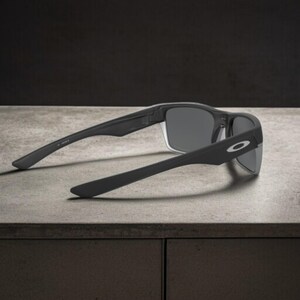 May include: Black sunglasses with gray lenses, featuring a sleek, modern design. The sunglasses have a matte finish and the Oakley logo is visible on the temple. The arms are extended, resting on a light gray surface.