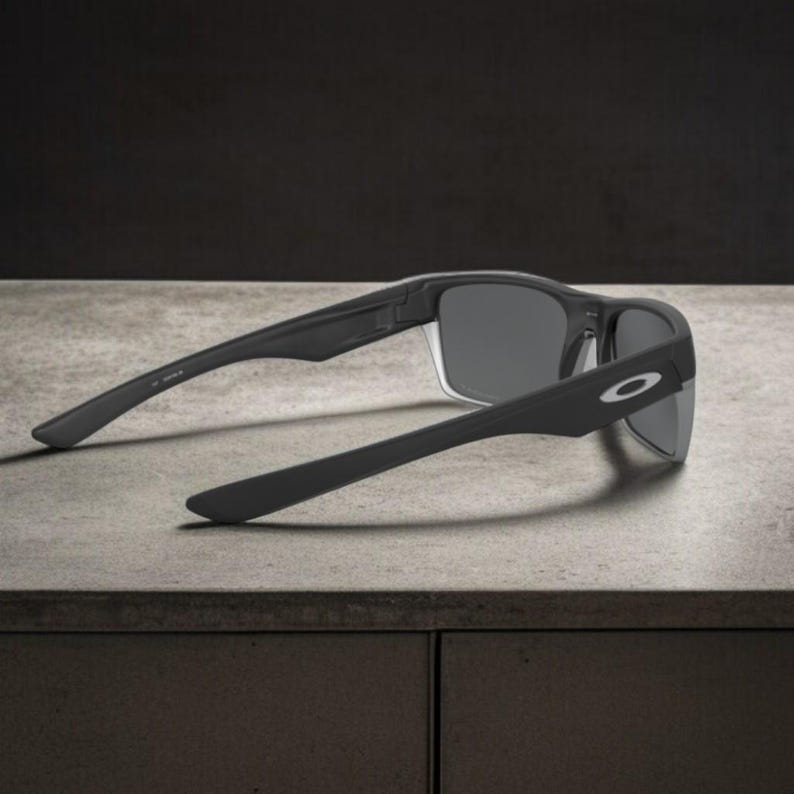 May include: Black sunglasses with gray lenses, featuring a sleek, modern design. The sunglasses have a matte finish and the Oakley logo is visible on the temple. The arms are extended, resting on a light gray surface.