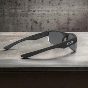 May include: Black sunglasses with gray lenses, featuring a sleek, modern design. The sunglasses have a matte finish and the Oakley logo is visible on the temple. The arms are extended, showcasing the full shape and style of the eyewear.