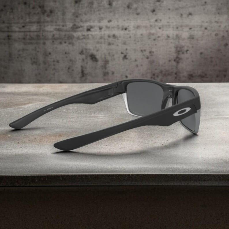 May include: Black sunglasses with gray lenses, featuring a sleek, modern design. The sunglasses have a matte finish and the Oakley logo is visible on the temple. The arms are extended, showcasing the full shape and style of the eyewear.