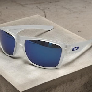 May include: Clear-framed sunglasses with blue mirrored lenses. The sunglasses have a rectangular shape and a small blue logo on the temples. The glasses are resting on a light-colored surface.