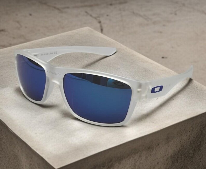 May include: Clear-framed sunglasses with blue mirrored lenses. The sunglasses have a rectangular shape and a small blue logo on the temples. The glasses are resting on a light-colored surface.