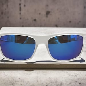 May include: White-framed sunglasses with vibrant blue mirrored lenses. The sunglasses are rectangular in shape and rest on a light-colored surface. The lenses reflect the surrounding environment, adding a dynamic element to the design.