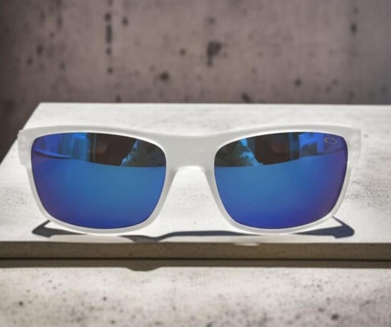 May include: White-framed sunglasses with vibrant blue mirrored lenses. The sunglasses are rectangular in shape and rest on a light-colored surface. The lenses reflect the surrounding environment, adding a dynamic element to the design.