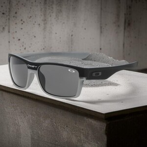 May include: Black and gray sunglasses with the Oakley logo on the temple. The sunglasses have a rectangular frame with gray lenses and a gray top bar. The sunglasses are resting on a concrete surface.