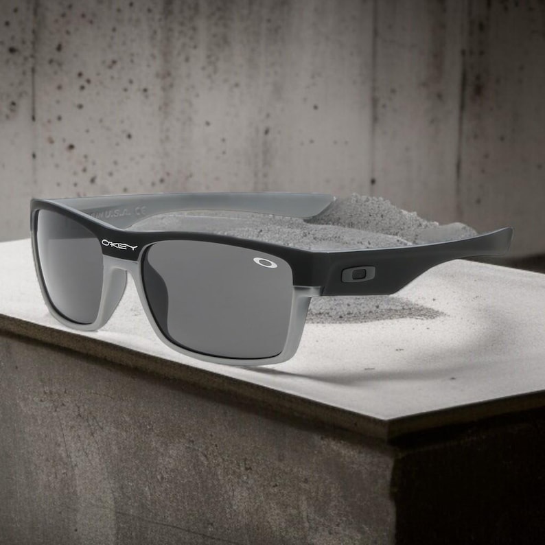 May include: Black and gray sunglasses with the Oakley logo on the temple. The sunglasses have a rectangular frame with gray lenses and a gray top bar. The sunglasses are resting on a concrete surface.