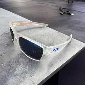 May include: Clear-framed sunglasses with blue lenses. The sunglasses have a rectangular shape and a white frame with a blue logo on the temple. The lenses reflect the surrounding environment.