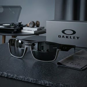May include: Black Oakley sunglasses with a gray frame and dark lenses. The sunglasses are displayed on a dark surface with a cleaning cloth and a box with the Oakley logo. A turntable and headphones are in the background.