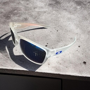 May include: White-framed sunglasses with blue lenses. The sunglasses have a sporty design with a blue logo on the temple. The lenses are dark, and the frame is a translucent white color. The sunglasses are resting on a gray surface.
