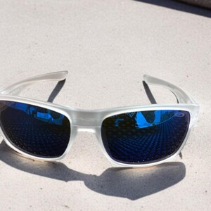 May include: White-framed sunglasses with blue mirrored lenses. The sunglasses are positioned on a light-colored surface, casting a shadow. The lenses reflect the surrounding environment, creating a vibrant visual effect.