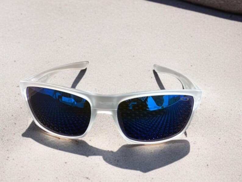 May include: White-framed sunglasses with blue mirrored lenses. The sunglasses are positioned on a light-colored surface, casting a shadow. The lenses reflect the surrounding environment, creating a vibrant visual effect.