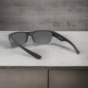 May include: Black rectangular sunglasses with gray lenses. The sunglasses have a clear frame around the lenses and black arms. The text "TWINFACE" is printed on the right arm.
