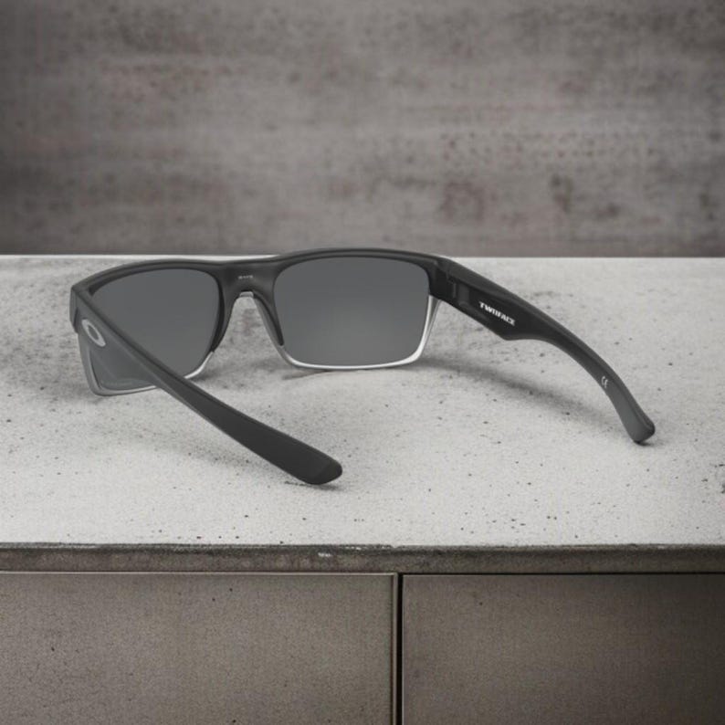 May include: Black rectangular sunglasses with gray lenses. The sunglasses have a clear frame around the lenses and black arms. The text "TWINFACE" is printed on the right arm.