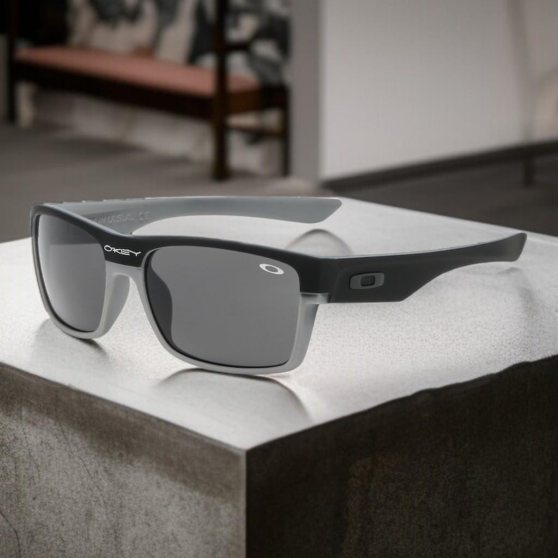 May include: Black and gray Oakley sunglasses with a rectangular frame. The sunglasses have dark gray lenses and a gray frame that contrasts with the black arms. The Oakley logo is visible on the frame.