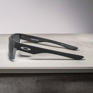 May include: Black sunglasses with gray lenses, featuring the word "TWINFACE" on the temple. The sunglasses have a matte finish and the Oakley logo is visible on the side. The glasses are resting on a light-colored surface.