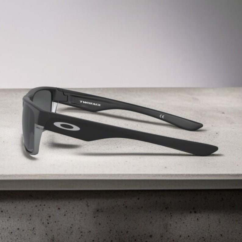 May include: Black sunglasses with gray lenses, featuring the word "TWINFACE" on the temple. The sunglasses have a matte finish and the Oakley logo is visible on the side. The glasses are resting on a light-colored surface.