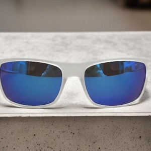 May include: White-framed sunglasses with vibrant blue mirrored lenses. The sunglasses have a rectangular shape and are resting on a light-colored surface. The lenses reflect the surrounding environment.