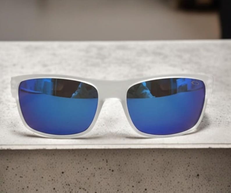 May include: White-framed sunglasses with vibrant blue mirrored lenses. The sunglasses have a rectangular shape and are resting on a light-colored surface. The lenses reflect the surrounding environment.