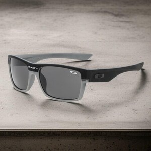 May include: Black and gray sunglasses with a rectangular frame. The sunglasses have gray lenses and gray arms. The Oakley logo is visible on the frame. The sunglasses are designed for outdoor use.