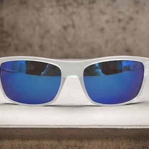 May include: White-framed sunglasses with vibrant blue lenses. The sunglasses have a rectangular shape and a modern design. The lenses reflect the surrounding environment, creating a mirrored effect. The sunglasses are displayed on a light-colored surface.
