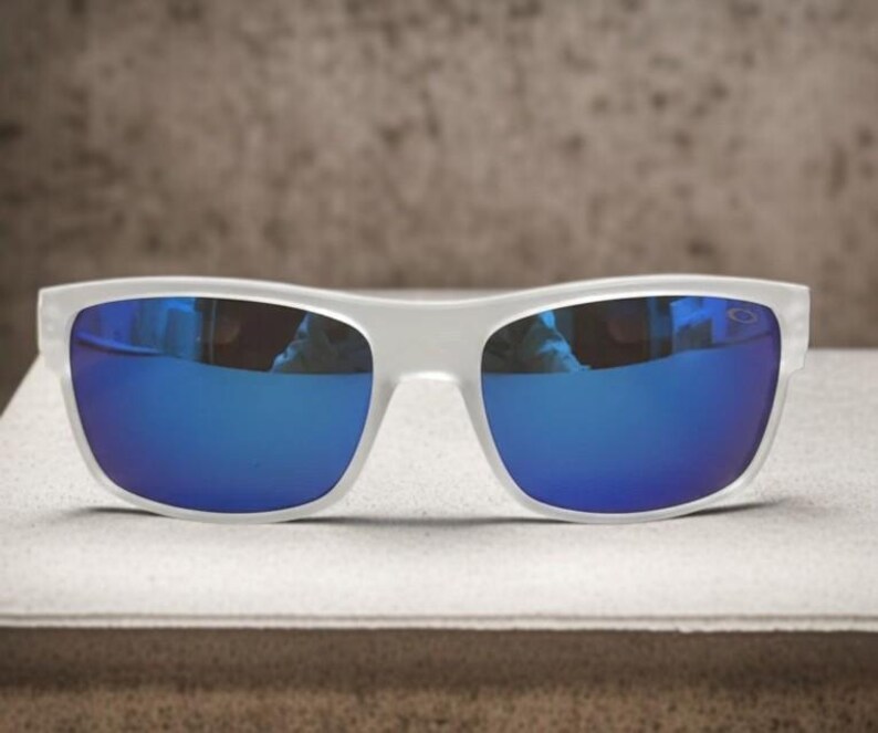 May include: White-framed sunglasses with vibrant blue lenses. The sunglasses have a rectangular shape and a modern design. The lenses reflect the surrounding environment, creating a mirrored effect. The sunglasses are displayed on a light-colored surface.