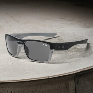 May include: Black and gray sunglasses with a rectangular frame. The sunglasses have a gray top bar and arms, with the brand name "Oakley" visible on the left lens. The lenses are a dark gray color.