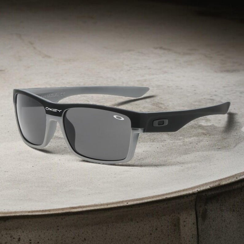 May include: Black and gray sunglasses with a rectangular frame. The sunglasses have a gray top bar and arms, with the brand name "Oakley" visible on the left lens. The lenses are a dark gray color.